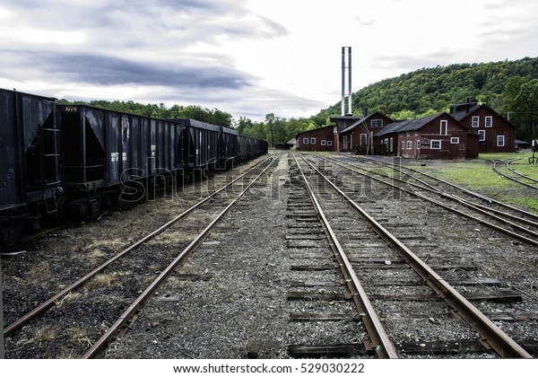 tracks running through old railroad yard next to rusting\
coal hoppers and maintenance shops with twin smoke stacks in rural\
area 