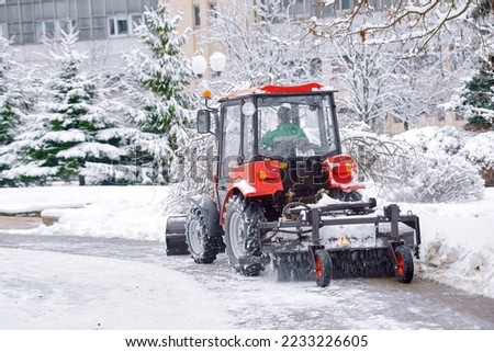 Tractor clear snow in park, machinery cleaning equipment. Snow removal red tractor clean walkway with plough sweeping brush. Tractor removing snow, cleaning sidewalk from snow in snowy winter