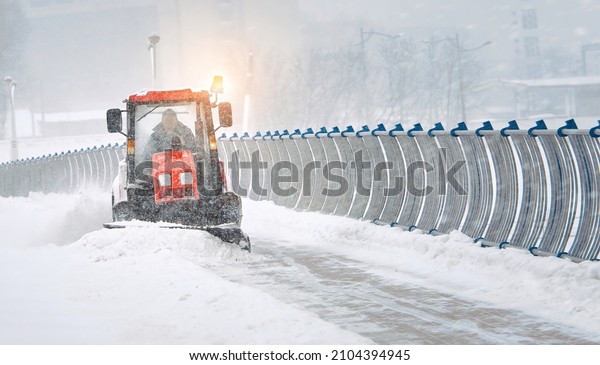 Tractor clear snow from pedestrian zone on car\
bridge, sidewalk snow management in the city during blizzard. Snow\
plow service clearing road from snow during snowstorm. Road\
maintenance in winter