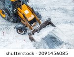 Tractor loader machine uploading dirty snow into dump truck. Cleaning city street, removing snow and ice after heavy snowfalls and blizzard. Snowplow outdoors clean pavement sidewalk road driveway