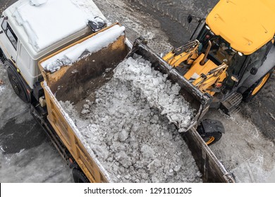 Tractor loader machine uploading dirty snow into dump truck. Cleaning city street, removing snow and ice after heavy snowfalls and blizzard