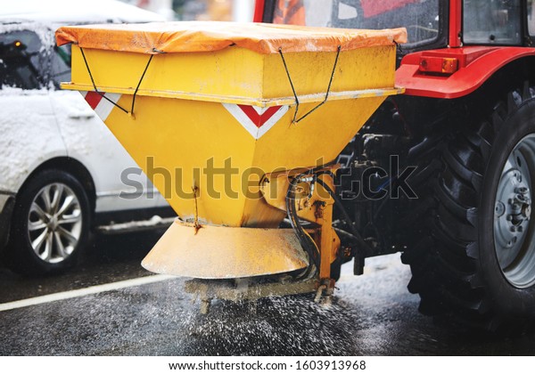 Tractor with mounted salt spreader, road maintenance\
- winter gritter vehicle.  Tractor de-icing street, spreading salt.\
Municipal service melting ice on streets. Diffuser of salt blend on\
road