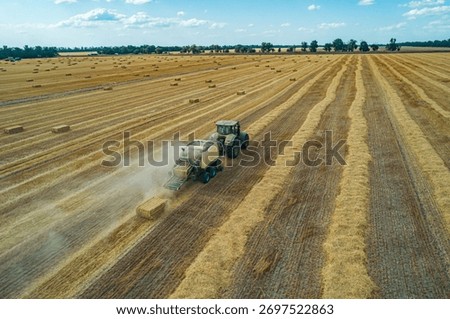 A tractor operates in a large field, creating hay bales during the summer harvest.