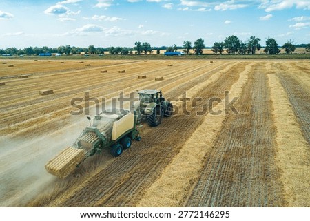 A tractor operates in a large field, creating hay bales during the summer harvest.