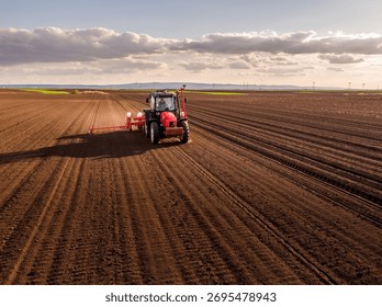 Tractor pulling a planter machine across a plowed field, seeding crops under a cloudy sky