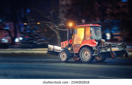 Tractor With Snow Plow And Rear Brush For Cleaning Sidewalks, Walkways And Roads Moving On City At Night, Emergency Road Maintenance At Winter Season. Tractor Cleaning Snow And Ice. Utility Equipment