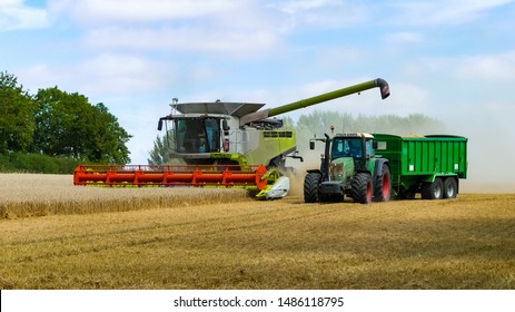 Tractor and trailer moves alongside a working combine harvester for it discharge grain from it's uploader in an English cornfield. Dust clouds behind. Landscape image with space for text. Oxfordshire. - Shutterstock ID 1486118795