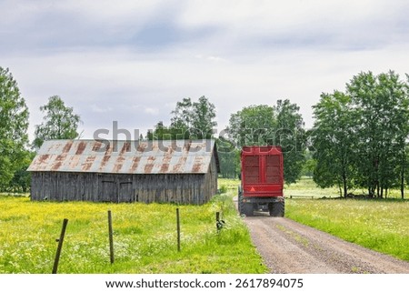 Tractor with a wagon driving on a gravel road by an old barn in a rural landscape