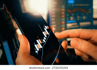 Trader analyzes financial data using mobile phone displaying live candlestick chart. Close-up view of hands monitoring stock market investments or cryptocurrency exchange rates. Background screens show additional financial graphs and metrics. Focus on technical analysis rapid decision making and modern mobile trading platforms.
