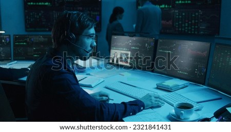 Trader with pen in hand works at computer with displayed real-time stocks. Colleagues analyze exchange market charts on big screens at background. Office illuminated by blue light. Investment concept.