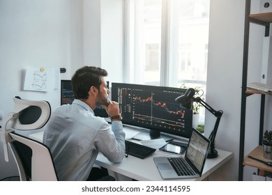 Trading on world markets. Young stock market broker analyzing data and graphs on multiple computer screens while sitting in modern office