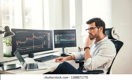 Trading online. Successful and young bearded trader in eyeglasses and formal wear working with laptop while sitting in his office in front of computer screens with trading charts - Shutterstock ID 1388871770