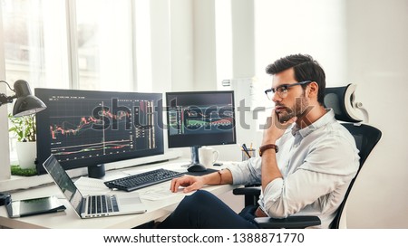 Trading online. Successful and young bearded trader in eyeglasses and formal wear working with laptop while sitting in his office in front of computer screens with trading charts