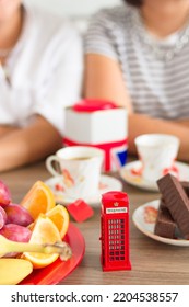 Traditional Afternoon Tea Of British Ceremony With Such Symbol Of Britishness As Toy Telephone Box, Defocused Faceless Women On Background, Front Focus, Vertical Image