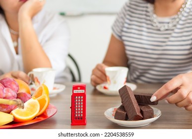 Traditional Afternoon Tea Of British Ceremony With Such Symbol Of Britishness As Toy Telephone Box, Defocused Faceless Women On Background, Front Focus