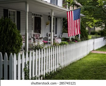 A Traditional American Home With A White Picket Fence And Flag Waving In A Small Town.