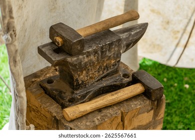 Traditional anvil with wooden hammer, set on a tree stump under a tent. Retro blacksmith tools used in crafts and metalworking. Photo taken outdoors, against a background of white canvas and green gra