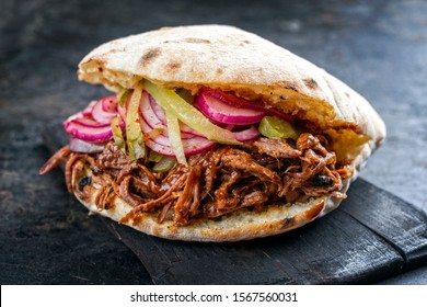 Traditional Barbecue Pulled Beef Burger Offered With Carolina BBQ Sauce, Gherkin And Onion Rings As Closeup On A Burnt Wooden Board 