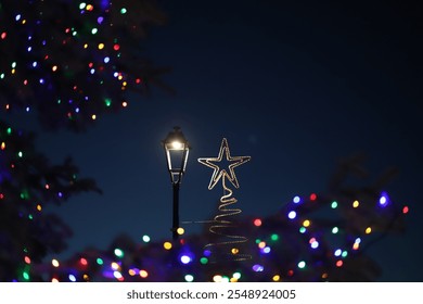 Traditional black street lamp post, with lit white bulb and  light rope in the shape of a Christmas Tree with a Star on top, backed by a dark night sky and surrounded with multicoloured fairy lights. - Powered by Shutterstock