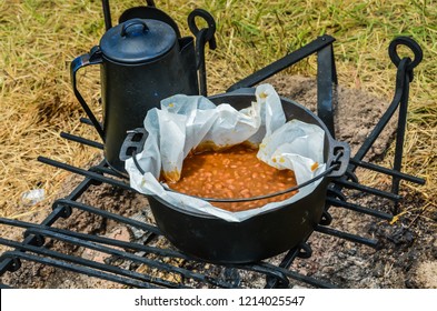 Traditional Boston Baked Beans cooked over hot coal in a black cast iron pot, with a kettle on the side. - Powered by Shutterstock