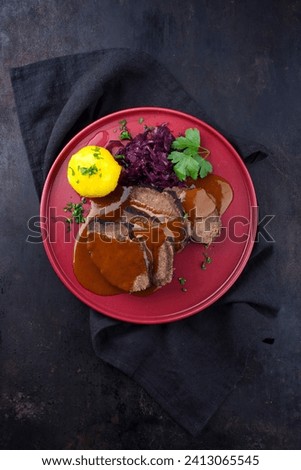 Traditional braised marinated German Sauerbraten from beef with blue kraut and potato dumplings in spicy dark beer sauce served as top view in a Nordic design plate with copy space