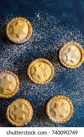 Traditional British Christmas Pastry Home Baked Mince Pies with Apple Raisins Nuts Filling Scattered on Ice Sugar Dusted Dark Blue Background. Golden Shortcrust Powdered Top View Copy Space
