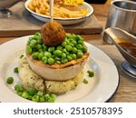 Traditional British pie served on a bed of mashed potatoes, topped with mushy peas and a battered meatball, accompanied by gravy on the side for a classic comfort meal.