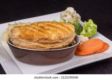 A Traditional British Pub Steak And Kidney Pot Pie With Flaky Crust In A Brown Casserole Dish With Vegetables And Mashed Potatoes On A Black Table