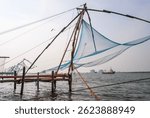 Traditional Chinese fishing net setup on the Arabian Sea coast in Kochi, Kerala, India. A local fisherman in a colorful wraparound skirt (lungi) is seen casting a handheld net near the shore, with lar