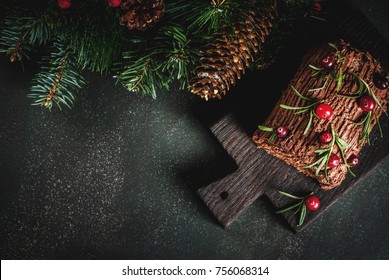 Traditional Christmas dessert, Christmas yule log cake with chocolate cream, cranberry and rosemary twigs. On a dark background with Christmas tree branches, gifts and decorations, copy space top view
