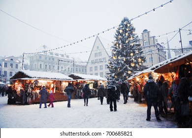 Traditional Christmas Market With A Christmas Tree, Lights, Booths And Warmly Dressed People At The Town Hall Square In Tallinn Old Town On A Snowy Day In Estonia.