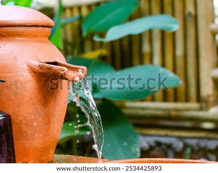 A traditional clay water pot with a spigot, surrounded by lush greenery. The pot is placed on a wooden base and a bottle of soap is placed beside it.