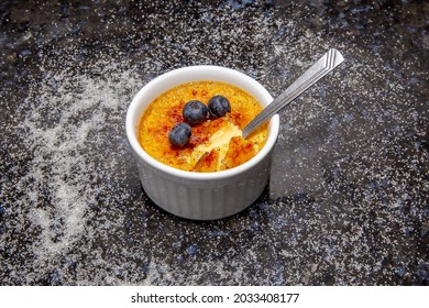 Traditional Crème Brûlée In Closeup Made With Vanilla Beans And Burnt With A Blowtorch. On A Dark Surface With Scattered Sugar And Decorated With Fresh Blueberries. Selective Focus