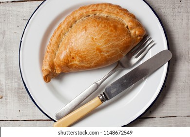A traditional Cornish pasty on a white enamel plate on a wooden table with a knife and fork