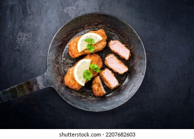 Traditional deep fried veal steak with lemon slices and herbs offered as top view in a rustic old wrought iron skillet with copy space 