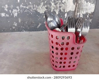 Traditional dining table setup featuring metal cutlery in a simple pink plastic holder, commonly found in Asian local diners or street food stalls. Stainless steel spoons and forks placed in a pink plastic cutlery holder on a dining table with a rustic concrete wall background. Spoons and forks organized in a pink plastic basket holder, close-up shot on a table against a weathered gray wall.