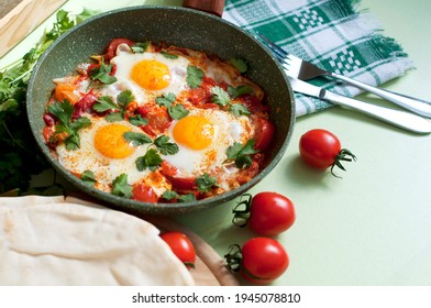 Traditional Dish Of Israeli Cuisine Shakshuka. Fried Eggs With Tomatoes, Peppers, Onions, Cilantro And Pita In A Pan On Light Green Background