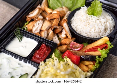 Traditional doner kebab in a black plastic container, sour cream, white boiled rice, roast meat, chiken, sauces, salad, cabbage and roasted vegetables on grey background