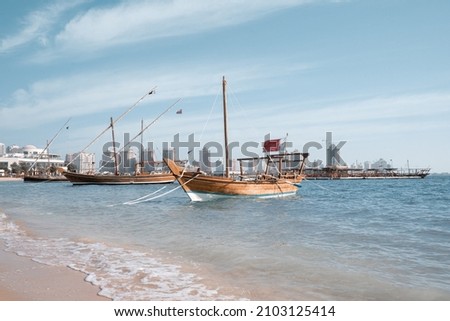 Traditional dow boat with Qatar flag harboured at a Katara beach, Doha, Qatar. Buildings and towers of Pearl Qatar in the background. 