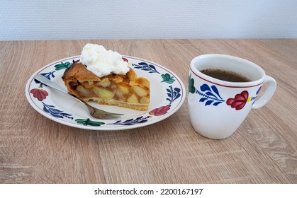 Traditional Dutch apple pie with whipped cream and a cup of coffee. Ceramics are in hand-painted farmer's pattern ('boerenbont') on a wooden table.