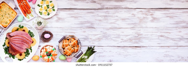 Traditional Easter Ham Dinner. Overhead View Corner Border On A White Wood Banner Background With Copy Space. Ham, Scalloped Potatoes, Vegetables, Eggs, Hot Cross Buns And Carrot Cake.