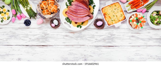 Traditional Easter ham dinner. Overhead view top border on a white wood banner background with copy space. Ham, scalloped potatoes, vegetables, eggs, hot cross buns and carrot cake.
