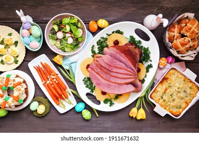 Traditional Easter ham dinner. Top down view table scene on a dark wood background. Ham, scalloped potatoes, vegetables, eggs, hot cross buns and carrot cake.