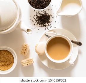 Traditional English Breakfast Black Tea With Milk -tea Leaves. Teacup, Teapot, Sugar And Milk Bowls, Macaroons Sweets On A White Background. Top View. Daylight. Close-up.