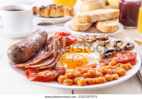 Traditional English Breakfast -\
plate with fried eggs, sausages, beans, mushrooms and bacon, cup of\
fresh coffee, croissants and orange juice on white\
background
