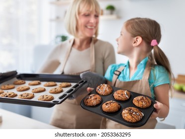 Traditional Family Recipe. Smiling mature woman and cheerful little girl holding tasty fresh baked cookies and muffins on trays while standing at dining table in kitchen, looking at each other