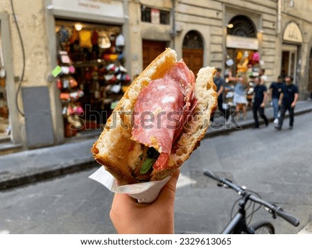 Traditional Florentine street food Sandwich with Schiacciata bread, Salami slices and rocket salad in a hand on blurry background of a street in Florence, Italy