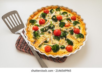 Traditional french pie. Quiche Loraine with mushrooms, broccoli, tomatoes and cheese. top view, in ceramic baking dish on  heat-resistant stand, pie spatula lies nearby