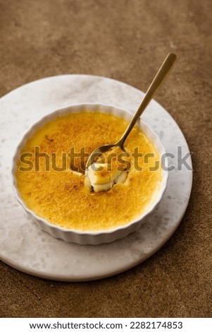 Traditional french portioned dessert creme brulee with creme, sugar and egg yolks. Burnt caramel crust. White ramekin on a marble stone stand. Brown background. Golden teaspoon with a creme brulee 