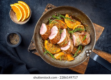 Traditional fried pork filet medaillons in with orange slices and herbs offered as top view in a rustic wrought iron skillet 